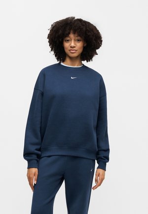 Kobieta z kręconymi włosami, ubrana w granatową bluzę i spodnie marki Nike, stojąca na jednolitym jasnym tle.
