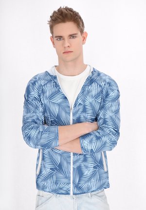 Blauwe vest met ritssluiting en een palmbladpatroon, voorzien van een capuchon, zijzakken en witte accenten. Draagt over een wit shirt.