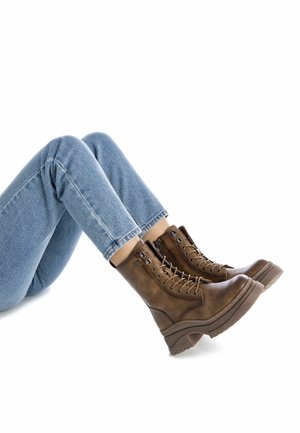 Botas de cuero marrón hasta el tobillo con diseño de cordones, suelas de plataforma gruesa y ojales de metal, combinadas con jeans azules enrollados.