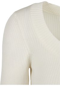 Gros plan d'un pull en maille côtelée couleur crème montrant le col en V et les détails de la couture aux épaules.
