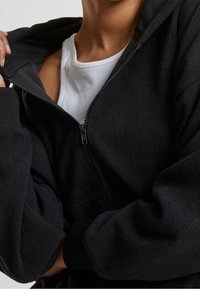 Person trägt eine strukturierte, schwarze, teilaufgeknöpfte Jacke mit Reißverschluss über einem weißen, gerippten Tanktop und hält mit einer Hand den Kragen der Jacke.