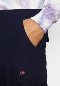 Main dans la poche d’un pantalon cargo bleu foncé avec broderie du drapeau norvégien, accompagnée d’une chemise à manches longues avec motif tie-dye violet et blanc.