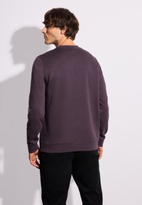Sudadera morada de manga larga y cuello redondo, que muestra una tela texturizada. El modelo se muestra de espaldas, vistiendo pantalones negros.