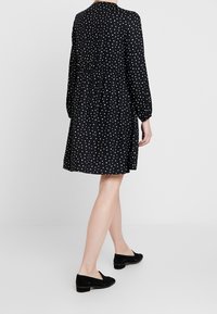 Robe noire à pois blancs, manches longues, coupe décontractée, taille froncée, longueur genoux, associée à des chaussures à enfiler en daim noir.