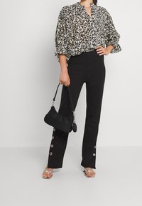 Schwarze Hochwasserhosen mit silbernen Knopfdetails, kombiniert mit einer locker sitzenden Bluse im Schwarz-Weiß-Muster und einer kleinen schwarzen Handtasche.