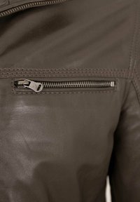 Braune Lederjacke mit einer Reißverschlusstasche auf der oberen linken Seite, metallischen Beschlägen, strukturiertem Oberflächenmaterial und kontrastierenden Nähten.