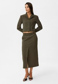 Olivengroene cropped blazer met knopen en revers, gecombineerd met een high-waisted, recht gesneden rok met een zijsplit. Zwarte open toe sandalen met hak.