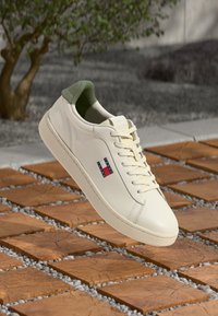 White leather sneaker with green heel accent, flat laces, and a logo tab; lightweight design with textured rubber sole.