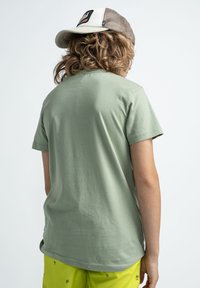 Světle zelené bavlněné tričko s krátkým rukávem, hladkou texturou a kulatým výstřihem, nošené s žlutými šortkami s malým vzorem palmových stromů.