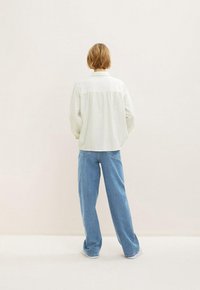 Camicia bianca a maniche lunghe con una vestibilità rilassata, abbinata a pantaloni larghi di jeans blu chiaro. Entrambi i capi sono mostrati di schiena.