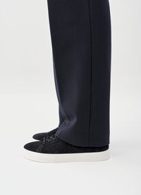Black suede sneakers with dark laces and white rubber soles, paired with tailored navy trousers, showing the shoe's profile and detail.