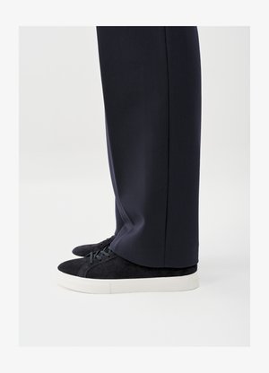 Mustad sametist tossud tumedate paelte ja valgete kummist talladega, mille juurde on kombineeritud eritellimusel valmistatud tumesinised püksid, näidates kinga külgvaadet ja detaile.