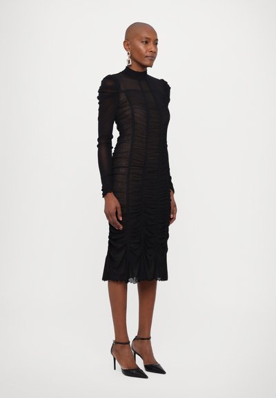 Rochie neagră cu mâneci lungi, transparente, textură plisată, gât înalt și siluetă de lungime medie. Asortată cu tocuri stiletto negre.