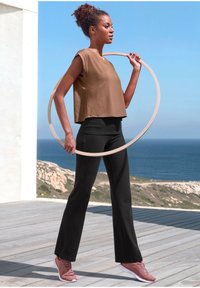 Braunes kurzärmeliges Oberteil kombiniert mit schwarzen Schlaghosen. Das Model hält einen Holzreifen und trägt rosa Sportschuhe. Im Hintergrund ist eine Küstenlinie zu sehen.