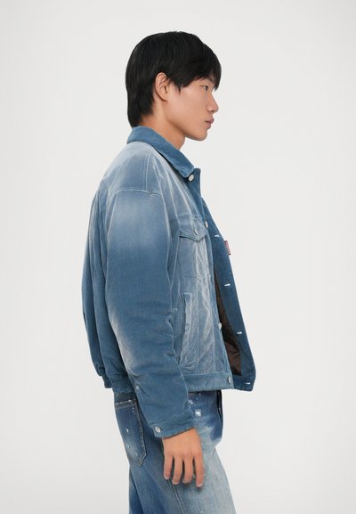Young man in faded blue denim jacket and distressed jeans facing right against plain background.