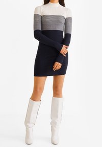 Femme portant une robe cintrée à blocs de couleur blanc, gris et bleu marine, associée à des bottes blanches montant jusqu'aux genoux, debout les bras croisés.