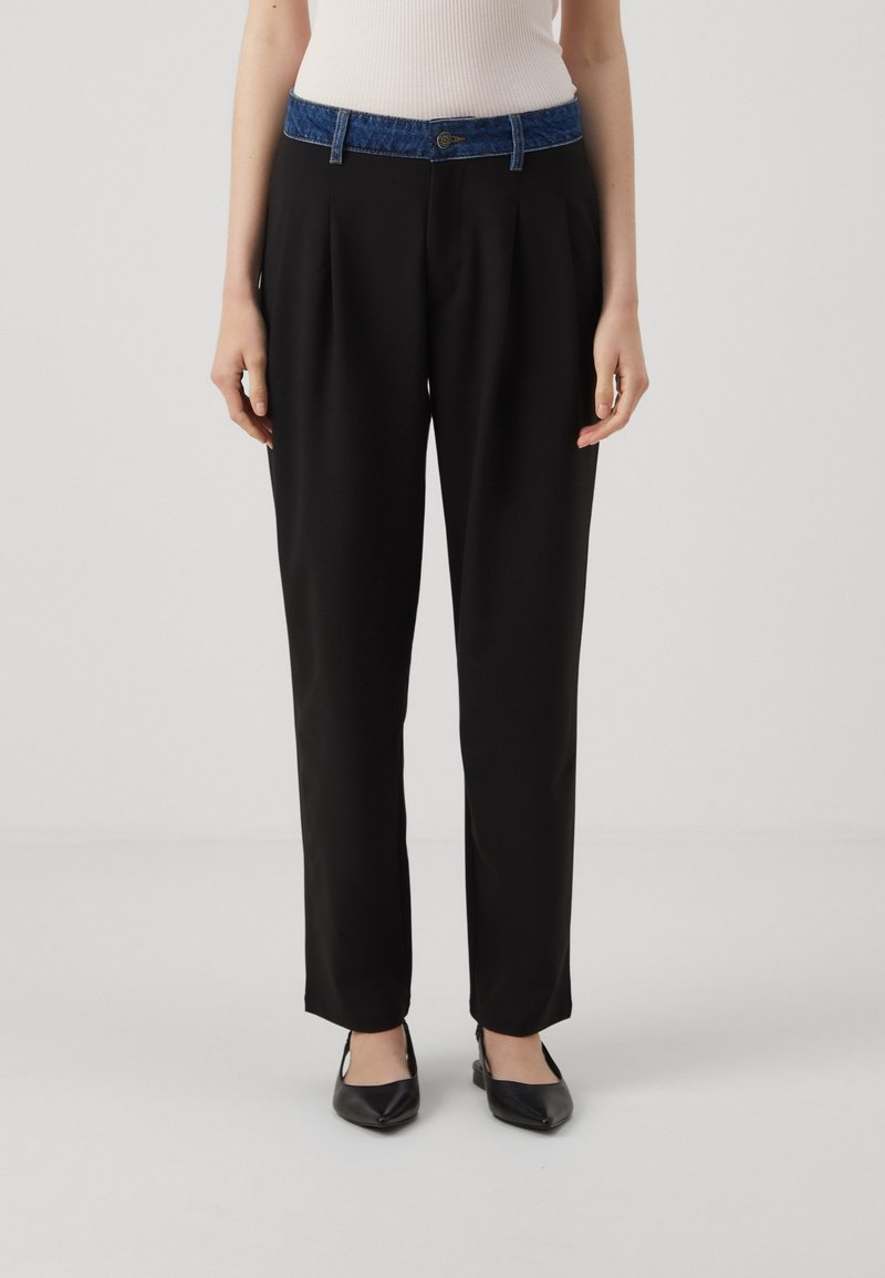 Femme portant un pantalon noir à jambes droites avec une ceinture en denim bleu et des chaussures noires à bouts pointus, debout devant un fond uni.