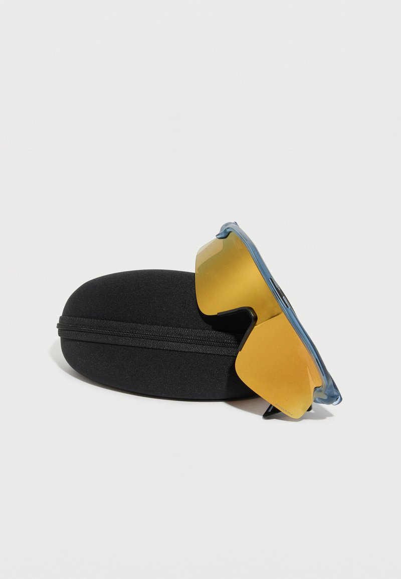 Lunettes de sport teintées de jaune avec des montures bleues posées contre un étui de protection noir zippé sur un fond blanc.