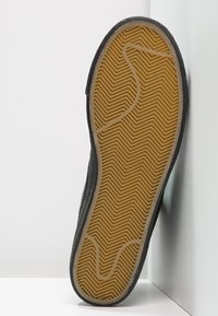 Schwarze Gummisohle mit Zickzackmuster. Sie verfügt über einen strukturierten Rand und eine hellfarbige, geriffelte Innenschicht für besseren Halt.