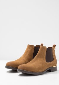 Un paio di stivali Chelsea in suede marrone con pannelli laterali elastici neri e tacco basso a stack su una superficie bianca.