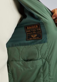 Zeleno prešito podstavljanje z oznako "RAIDER," ki podaja številko izdaje in oblikovanje. Ima stransko žep v usklajenem materialu.