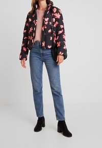 Schwarze Steppjacke mit rosa Blumenmuster, Reißverschluss vorne, hoher Kragen. Kombiniert mit hellblauen Jeans und schwarzen Stiefeln mit Absatz.