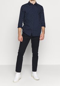 Camisa de botón azul marino con patrón de formas geométricas blancas, con mangas enrolladas de 3/4, combinada con pantalones negros ajustados y zapatillas blancas.