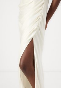 Elfenbenfärgad klänning med draperad textur, featuring en höga slits och slätt tyg som framhäver en elegant silhuett.