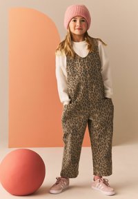 Pantalones de tirantes con estampado de leopardo en tonos marrón y negro, que presentan grandes bolsillos frontales. Combinados con un suéter blanco y un gorro de punto rosa, además de zapatillas rosas.