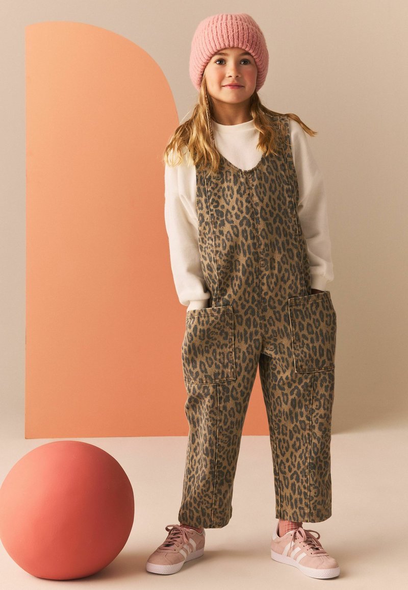 Pantalones de tirantes con estampado de leopardo en tonos marrón y negro, que presentan grandes bolsillos frontales. Combinados con un suéter blanco y un gorro de punto rosa, además de zapatillas rosas.