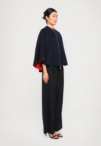 Cape marine avec doublure rouge, présentant une fermeture éclair à l'avant. Associé à un pantalon large plissé noir et des sandales à talons noires.