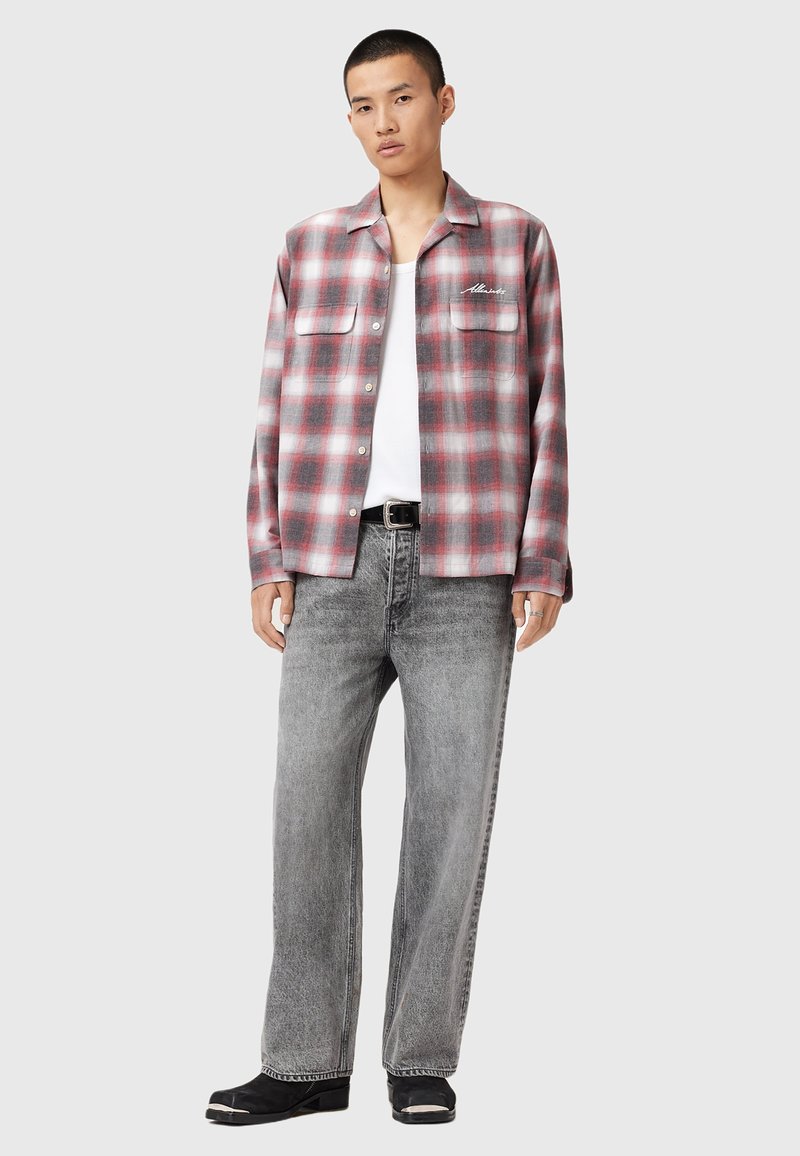 Homme portant une chemise à carreaux rouge et gris sur un haut blanc, un pantalon en denim large gris délavé, une ceinture noire et des chaussures noires avec des embouts en métal argenté.