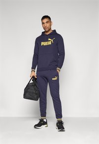 Felpa con cappuccio blu navy e pantaloni della tuta, con logo giallo "PUMA" e polsini a righe. Scarpe da Ginnastica nere e una piccola borsa nera.
