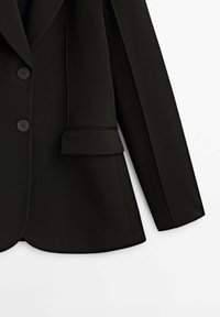 Blazer noir cintré avec deux boutons sur le devant, une poche à rabat côté droit et une manche longue sur fond blanc.