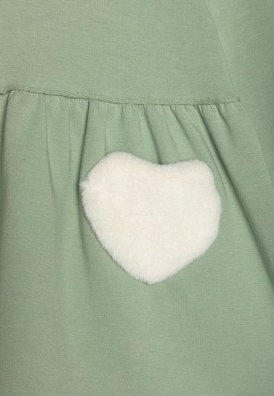 Zeleni tekstilni top z nabranim robom. Opremljen je z belim, puhastim, srčastim zakrpom na strani. Mehka tekstura, sproščen design.