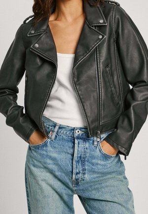 Femme portant une veste de motard en cuir noir sur un débardeur blanc et un jean bleu clair taille haute, les mains dans les poches.