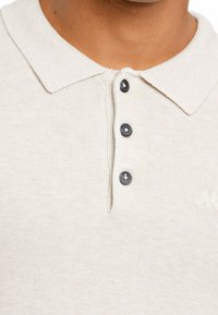 Beżowa koszulka polo z teksturowanej tkaniny, trzema granatowymi guzikami i haftowanym logo na lewej piersi. Klasyczny krój kołnierzyka.
