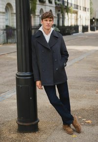 Young man in dark checkered coat and jeans leans against black lamppost on a quiet urban street with autumn leaves scattered.