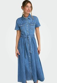 Femme portant une robe chemise en denim bleu à manches courtes avec des poches poitrine et une ceinture à la taille, regardant vers sa gauche.