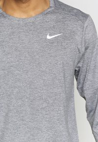 Camiseta atlética de manga larga gris, hecha de material texturizado; cuenta con un logo blanco de Nike Swoosh en la parte superior izquierda del pecho.