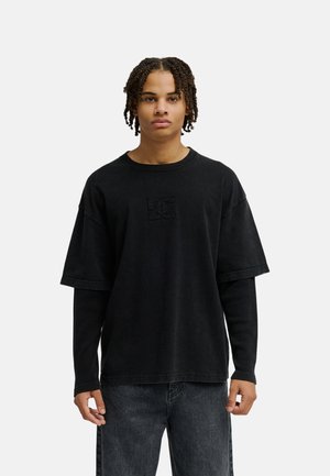 Junger Mann mit kurzen Dreadlocks, der ein geschichtetes schwarzes Langarm- und kurzärmeliges Shirt sowie dunkle Jeans trägt und vor einem weißen Hintergrund steht.