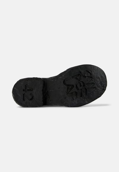 Suela de zapato de goma negra con una superficie texturizada, que presenta logotipos en relieve y talla 42. El diseño incluye una forma irregular y un patrón de profundidad en la banda de rodadura.