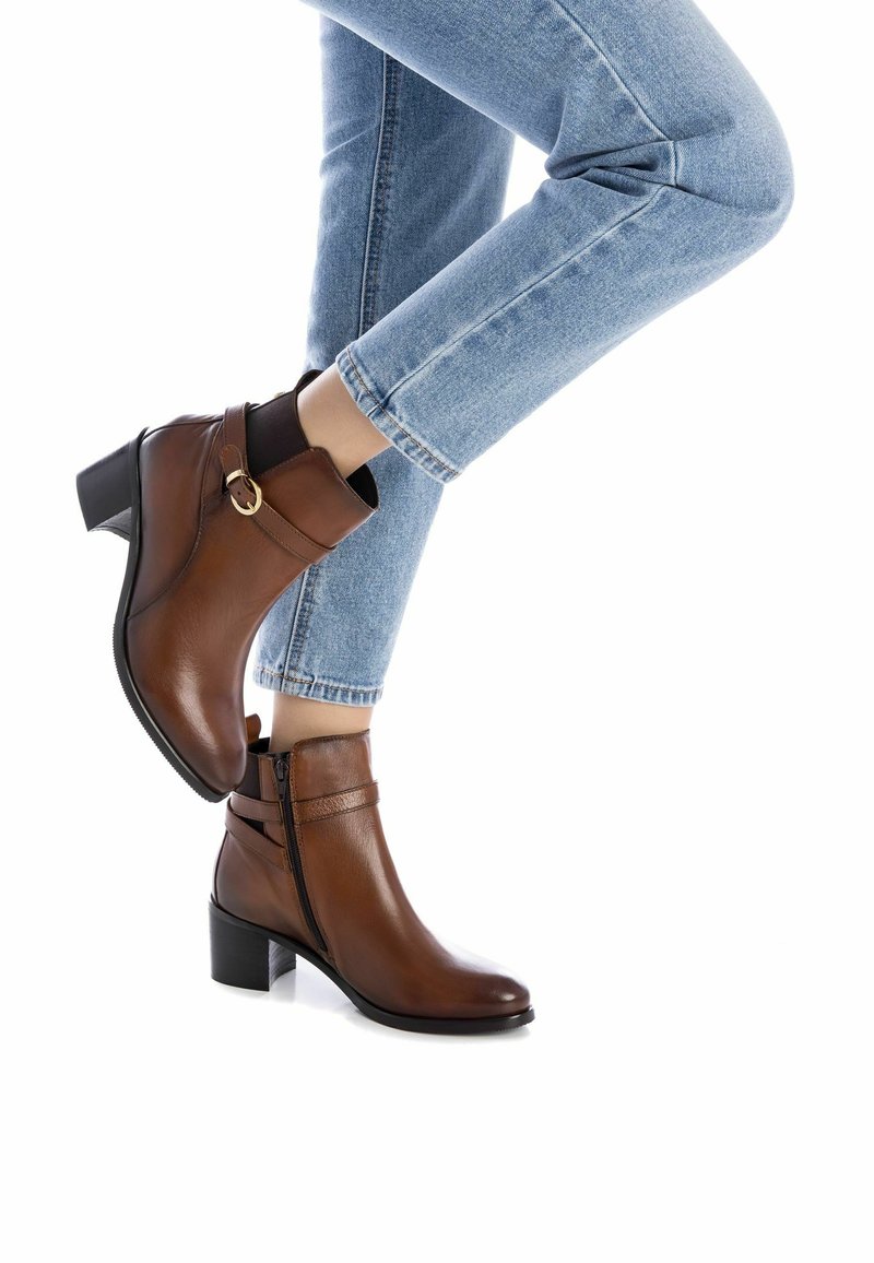 Botas de tobillo de cuero marrón con cremallera lateral, correa decorativa y un tacón de bloque medio. Llevadas con jeans claros con dobladillo.