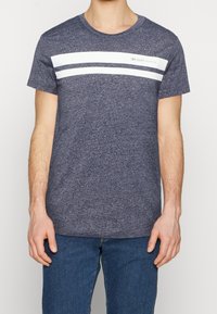 Mężczyzna ubrany w melanżową granatową koszulkę z dwoma białymi poziomymi paskami i napisem "DENIM TOM TAILOR", połączoną z niebieskimi dżinsami.