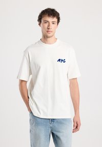 White cotton t-shirt with short sleeves and crew neck. Features blue embroidered "NYC" text on the left chest. Paired with light denim jeans.