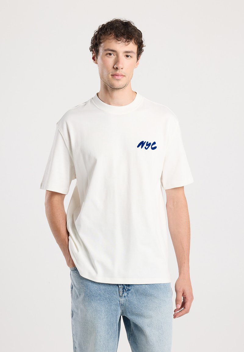 White cotton t-shirt with short sleeves and crew neck. Features blue embroidered "NYC" text on the left chest. Paired with light denim jeans.