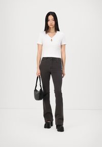 Top bianco a maniche corte con scollatura a chiave, abbinato a jeans svasati grigio scuro. Il modello tiene una borsa nera con texture e indossa scarpe nere chunky.