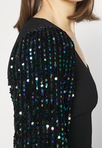 Détail aux épaules en sequins noirs sur un haut noir ajusté et côtelé, présentant un aspect texturé avec des accents colorés et réfléchissants.