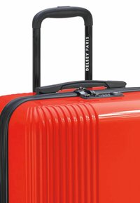 Valigia rigida rossa con scanalature verticali, zip nera e manico, branding stampato sul manico e un design compatto.