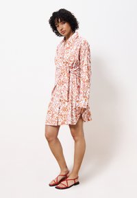 Robe à motif floral avec une base rose et des motifs orange, manches longues, taille ceinturée et un ourlet au genou. Assortie avec des sandales rouges.